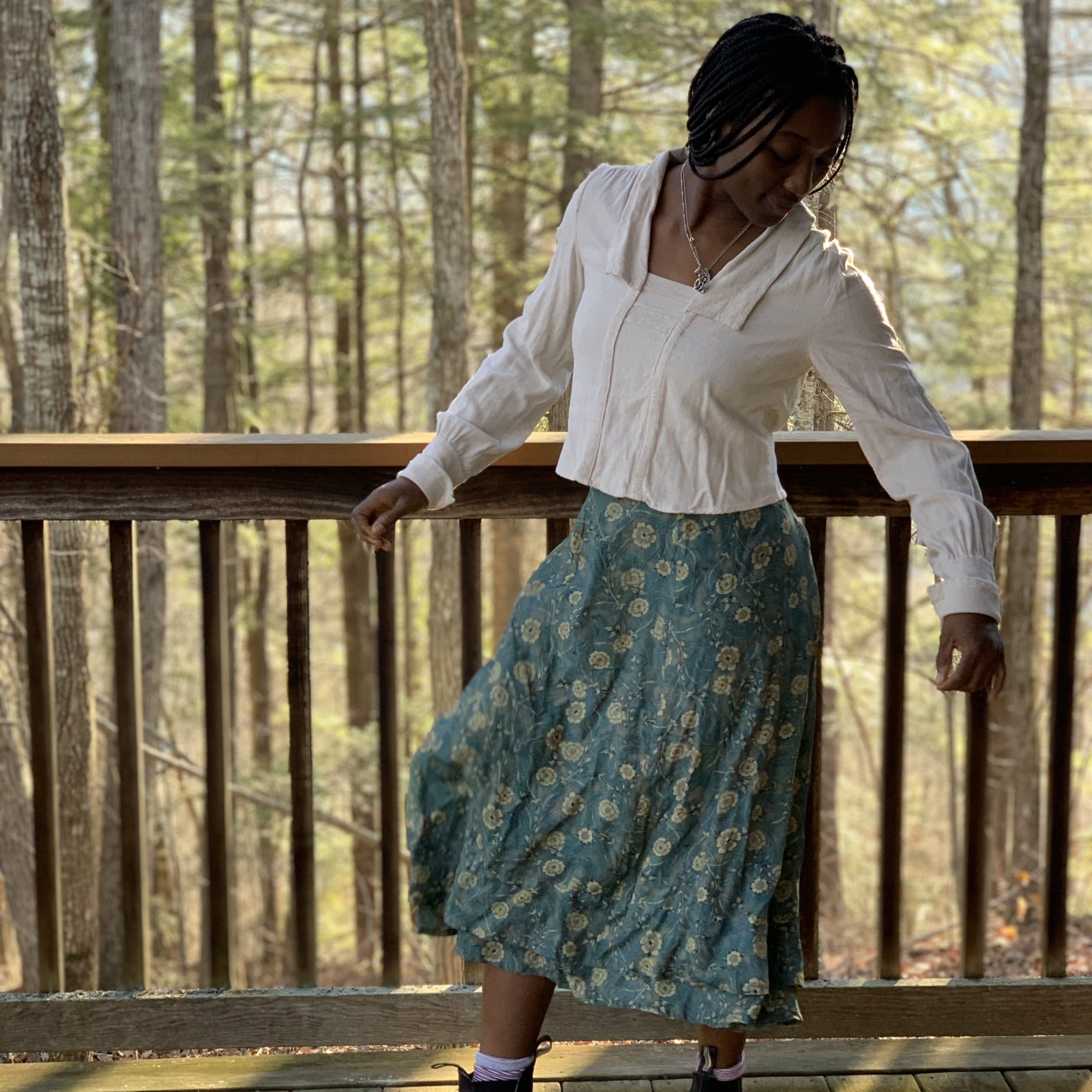 African American Woman twirling towards her right, wearing 210 armistice blouse in a light beige and  209 walking skirt in floral teal/taupe viscose fabric. On a porch outside.