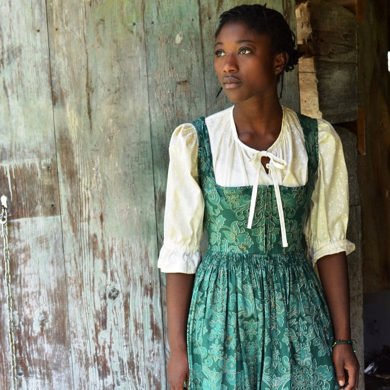Black woman wearing a green floral Dirndl and white blouse against an old barn door.