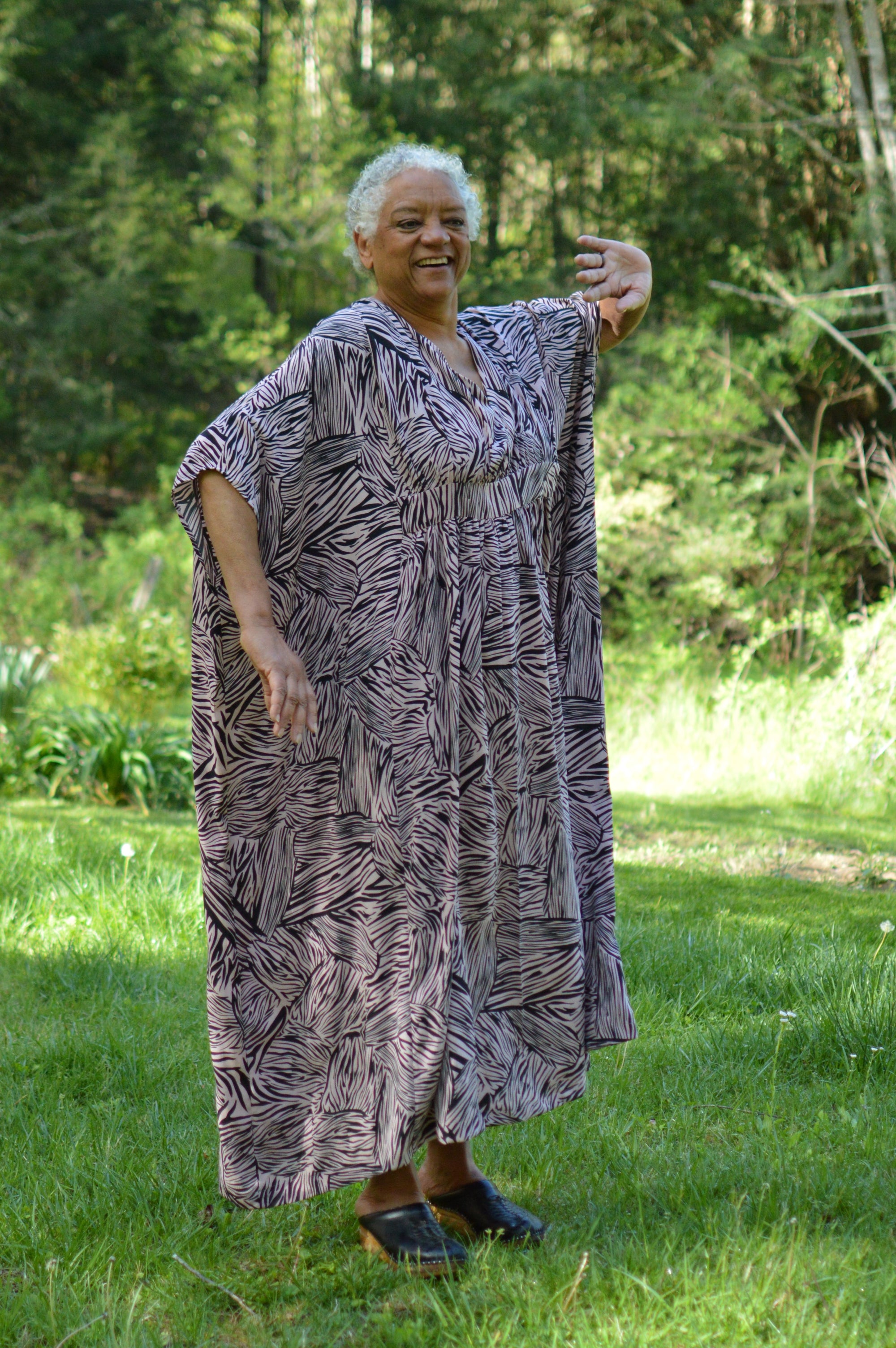Dancing older African American woman wearing Kaftan in a black and pink stripe like print with greenery in the background.