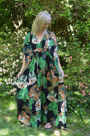 White woman wearing a tropical green and orange print kaftan standing out side in front of greenery and white and pink flowers.