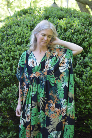 White woman wearing a tropical green and orange print kaftan standing out side in front of greenery with her hand on her neck.