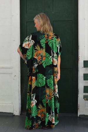 Back of white woman wearing a tropical green and orange print kaftan standing  in front of a green door.