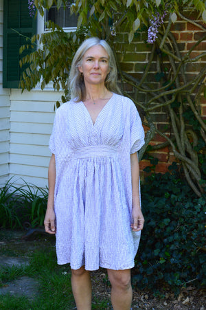 white woman wearing a knee length Kaftan in a lilac  and white stripe fabric standing in front of a house with greenery.