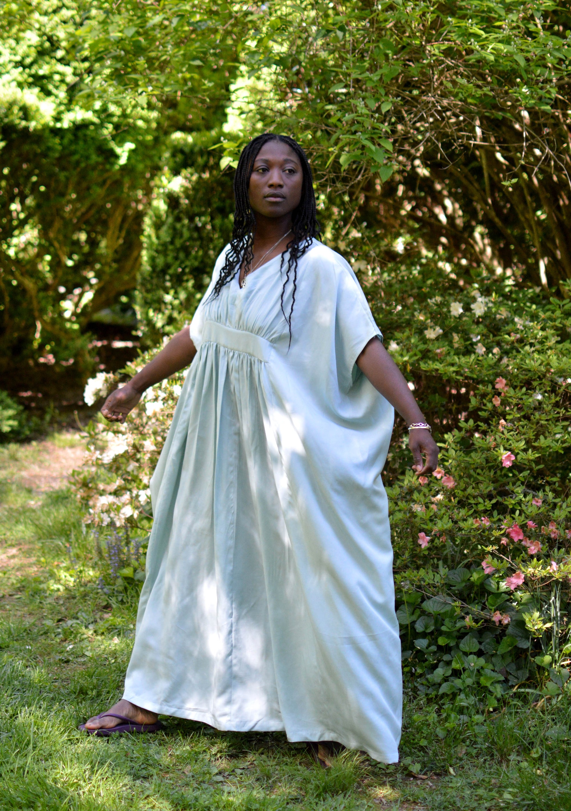 African American woman wearing a light aqua blue kaftan with arms out standing outside with a greenery background.