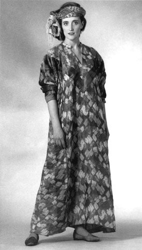 Woman wearing 105 Syrian Dress.  Dress is ankle length and sleeves are pushed up to below elbow.  fabric is an abstract geometric print.  Model has a scarf tied around head and is photographed with a white background.