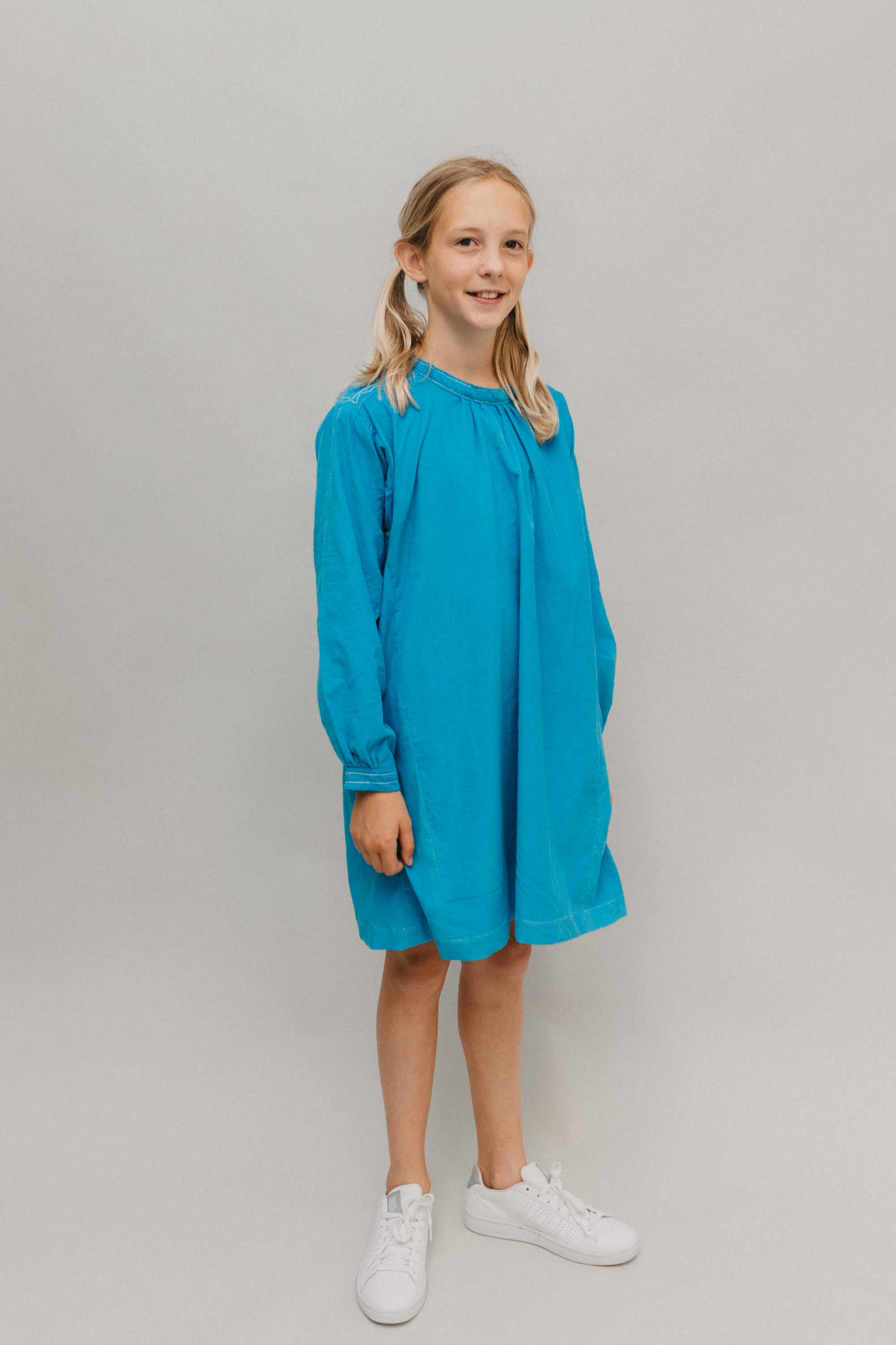Photo of young girl wearing a blue corduroy Smock.  Model is standing in front of a white background looking towards camera.  