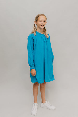 Photo of young girl wearing a blue corduroy Smock.  Model is standing in front of a white background looking towards camera.  