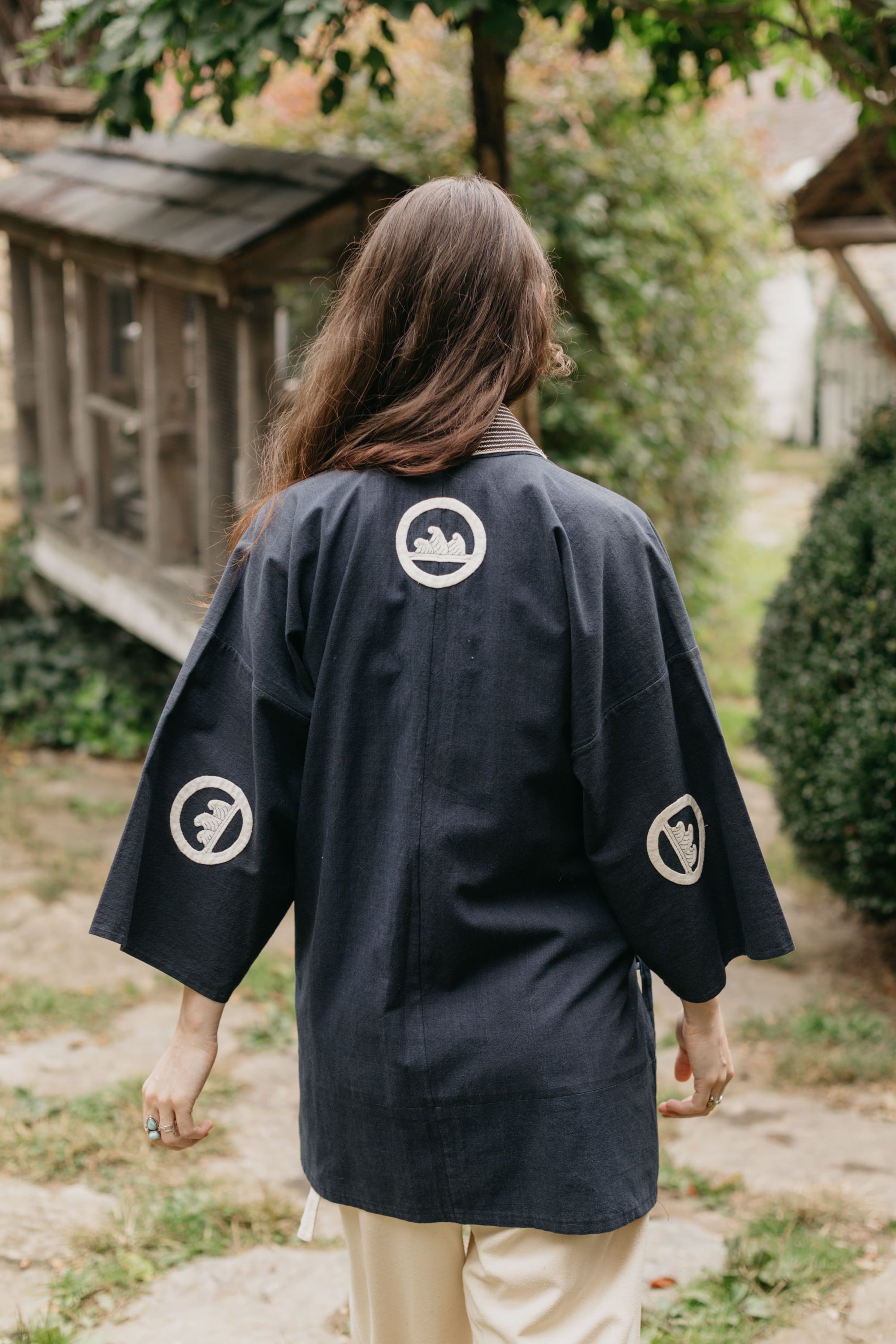 Japanese American woman wearing a navy Hippari with embroidery. Back View, outside by shrubs.  Woman is walking away.