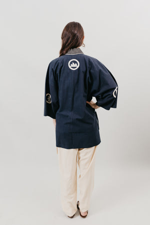 Japanese American woman wearing a navy Hippari with embroidery. Back View, with cream colored pants on a white background.