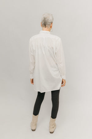 Woman standing in front of light grey background wearing a white Croatian shirt as a tunic with black leggings and boots.  Her back is facing.