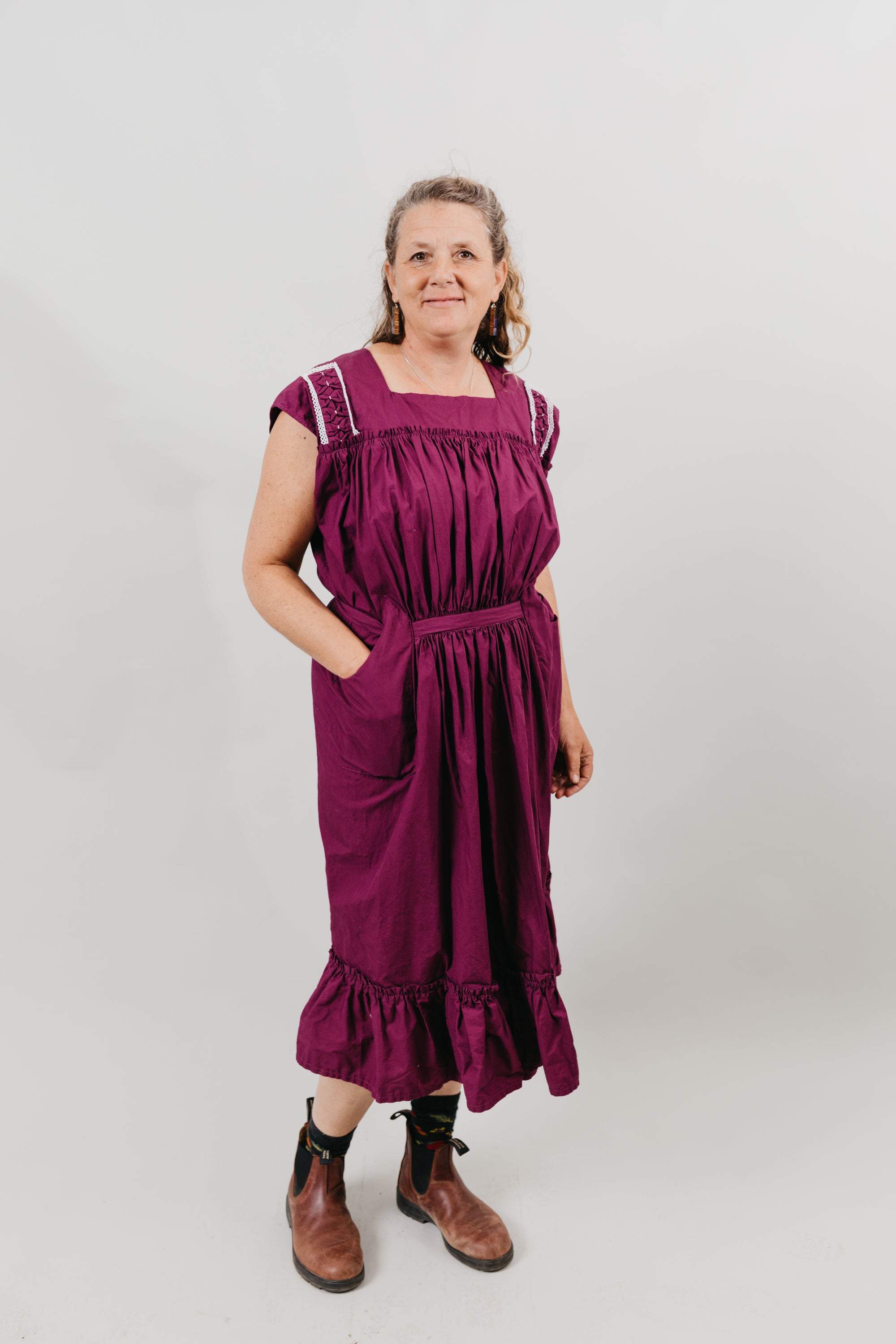 White woman wearing a purple sleeveless dress with embroidery at the shoulder yokes.  Standing in front of a white background.  Dress has pockets and is belted at the waist and has a flounce at the bottom.