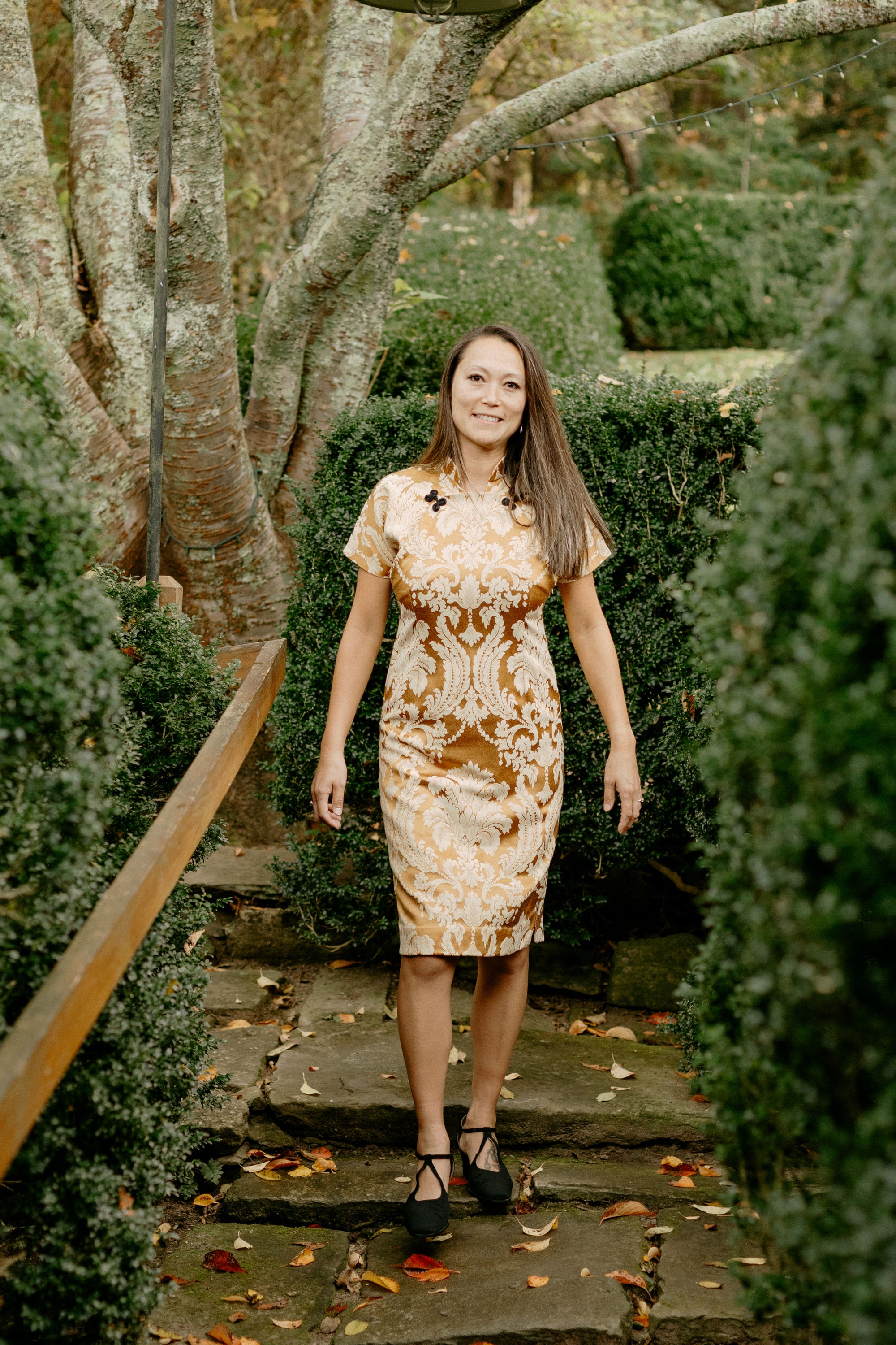 Woman walking down stone steps by shrubs wearing a knee-length gold brocade cheongsam.
