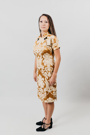 Asian woman wearing a gold brocade knee-length cheogsam, standing sideways in front of a white background.