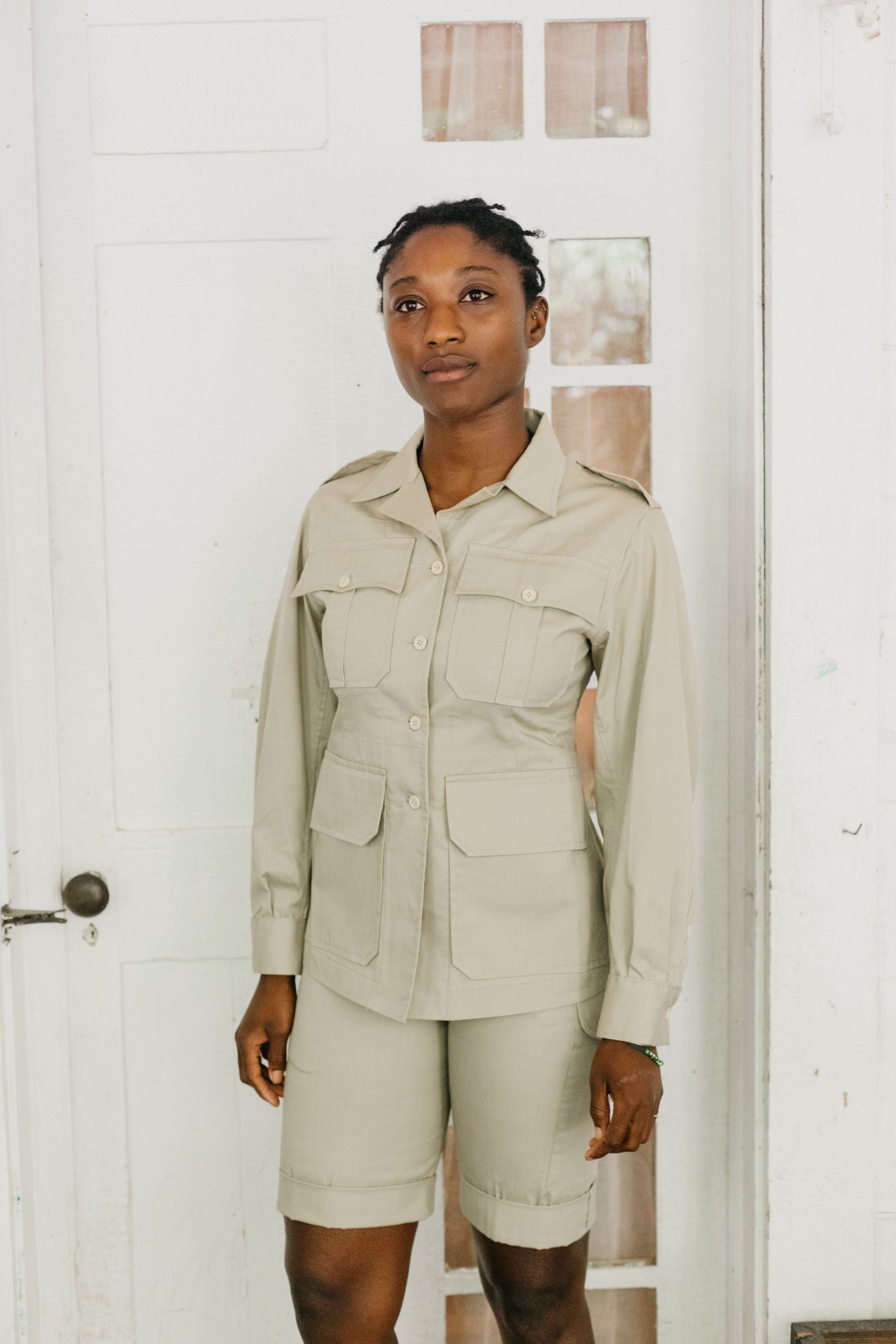 Black woman wearing long sleeve khaki  bush shirt and shorts in front of a white door.