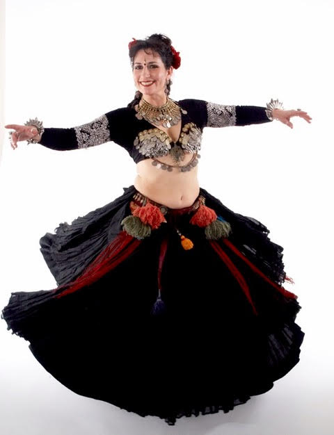 Woman wearing a full black skirt, tasseled belt, cropped long sleeve black choli and coin bra.  She is spinning and the skirt is flaring out and her hands are out to the side.  She is a belly dancer.