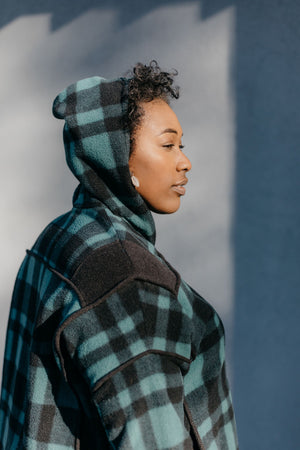 Close up of Black woman standing outside in front of a grey wall wearing a green and black plaid siberian parka. The hood is up and she is facing to the right.
