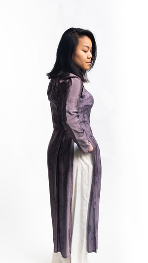 Side view of Vietnamese American woman wearing a purple silk Ao Dai tunic with white pants and sandals in front of white backdrop.