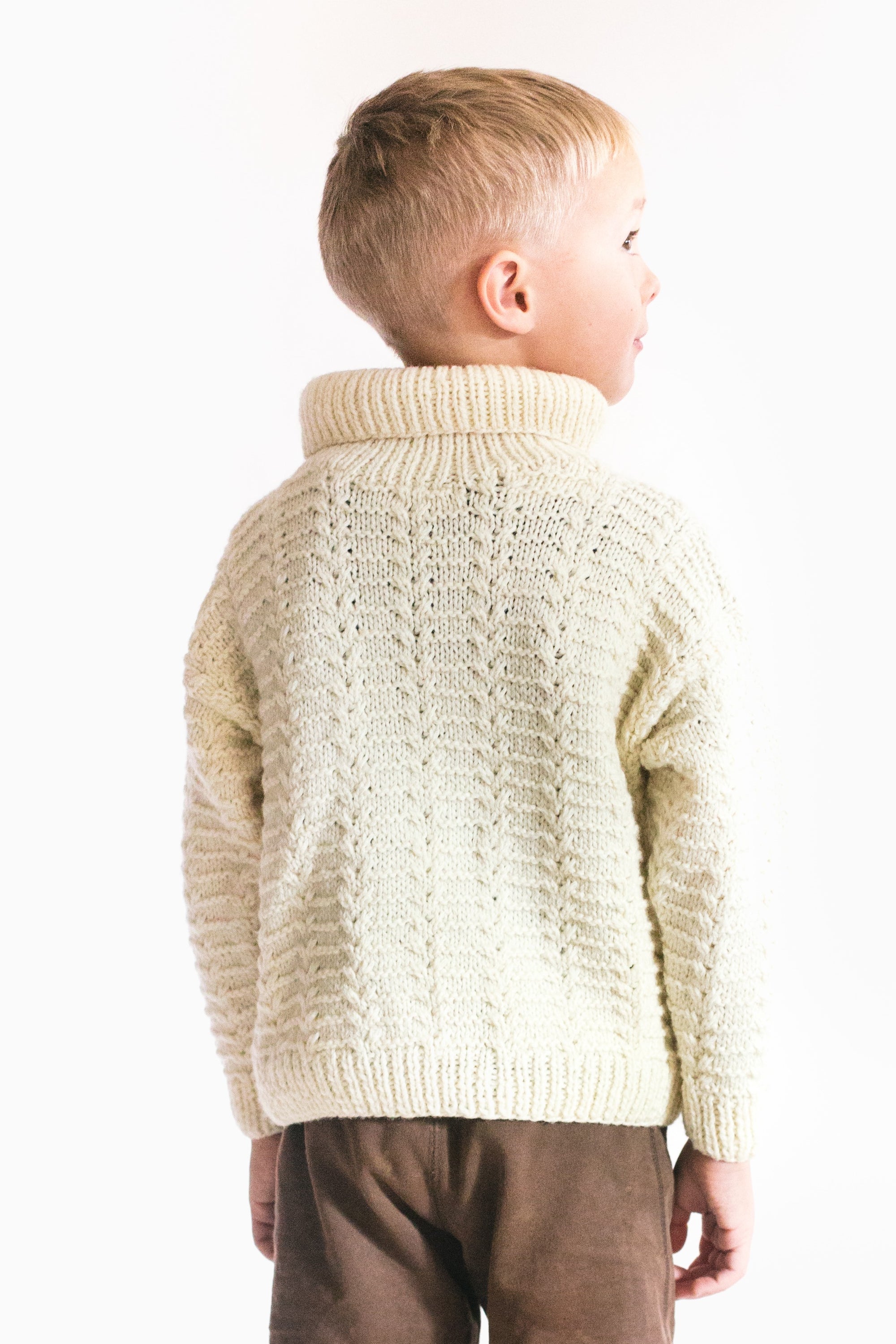 Boy wearing a cream colored hand knit sweater with a rolled neck, and brown shorts.  Standing in front of a white backdrop.