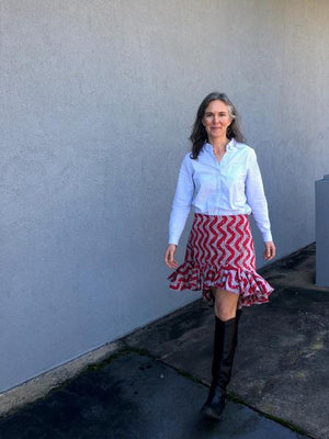 Woman standing in front of a grey wall outside, wearing a buttondown shirt with the Flamenco practice skirt and black boots.  Skirt is red and white ankara fabric and is above the knee in the front and top of calf in the back with a ruffled flounce.  Woman is walking. 