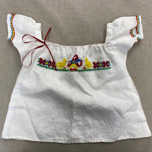 Flat lay of the Mexican Baby Dress in white linen with red, blue, yellow, and green embroidery on the yoke and sleeves.