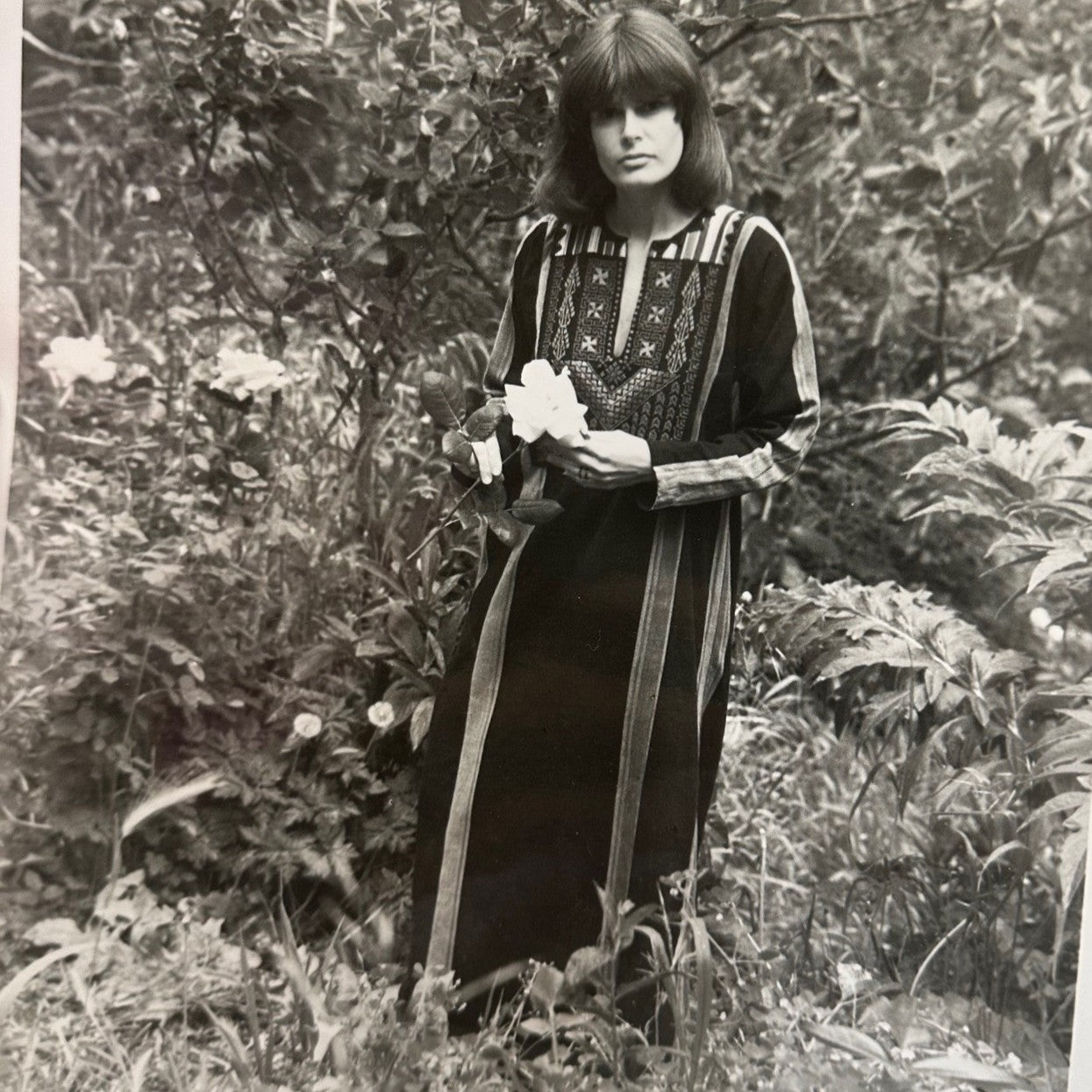 Black and white photo of a woman standing outside holding a flower and wearing an embroidered gaza dress