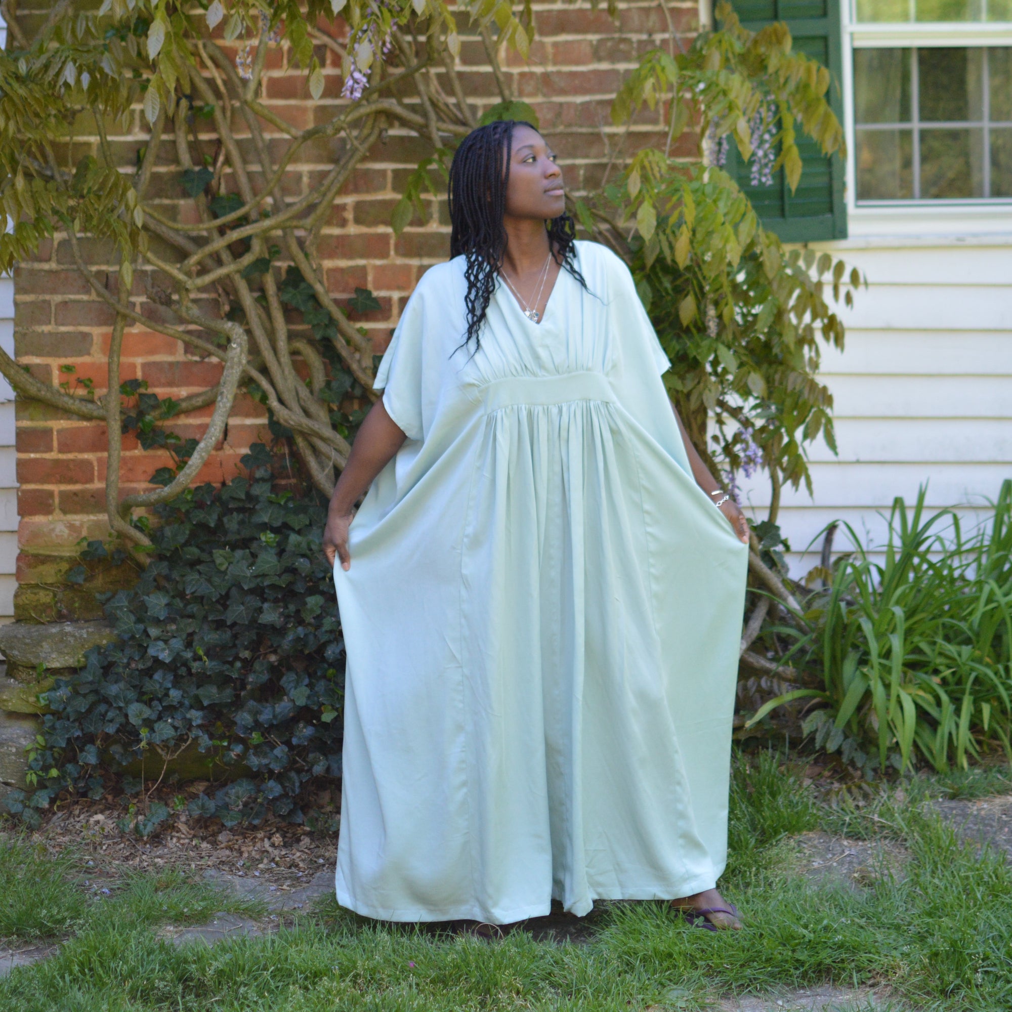 Black woman with long hair standing outside in front of a brick chimney and a white house with wisteria growing on it, wearing a light blue kaftan dress.