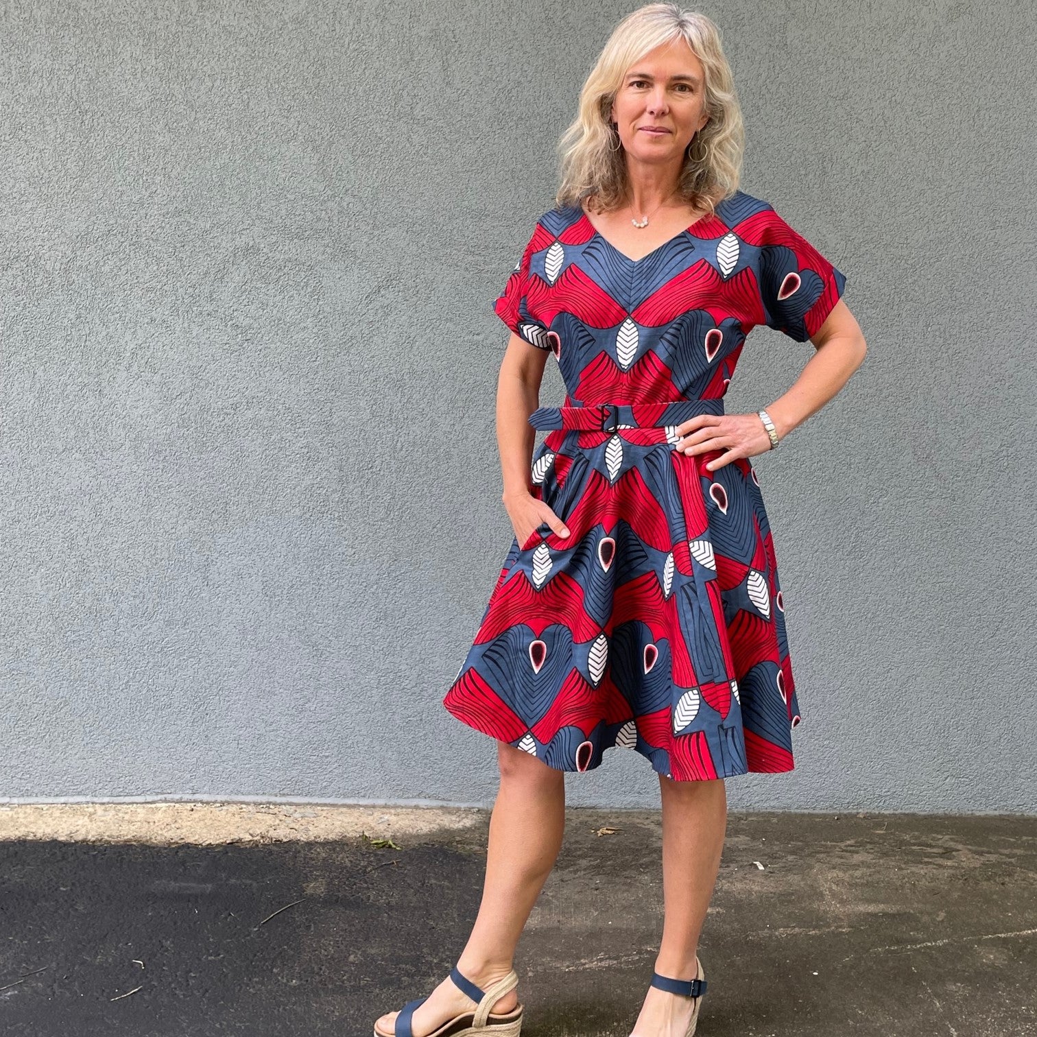 Woman standing in front of a grey wall wearing a blue and red fit and flair dress.  Left hand on hip, right hand in pocket.