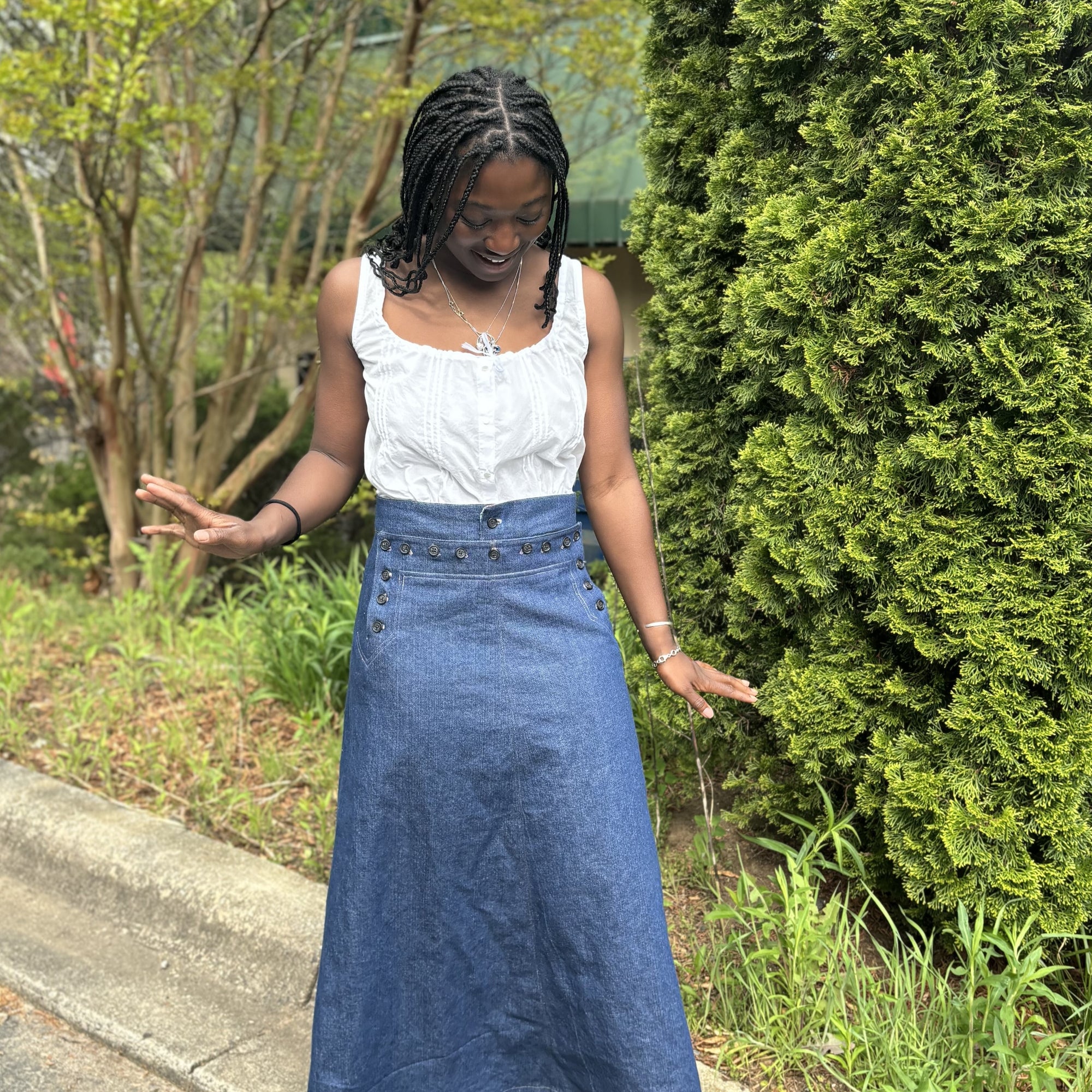 African American woman standing looking down wearing a long denim sailor skirt with a sleeveless white blouse with greenery in the background.