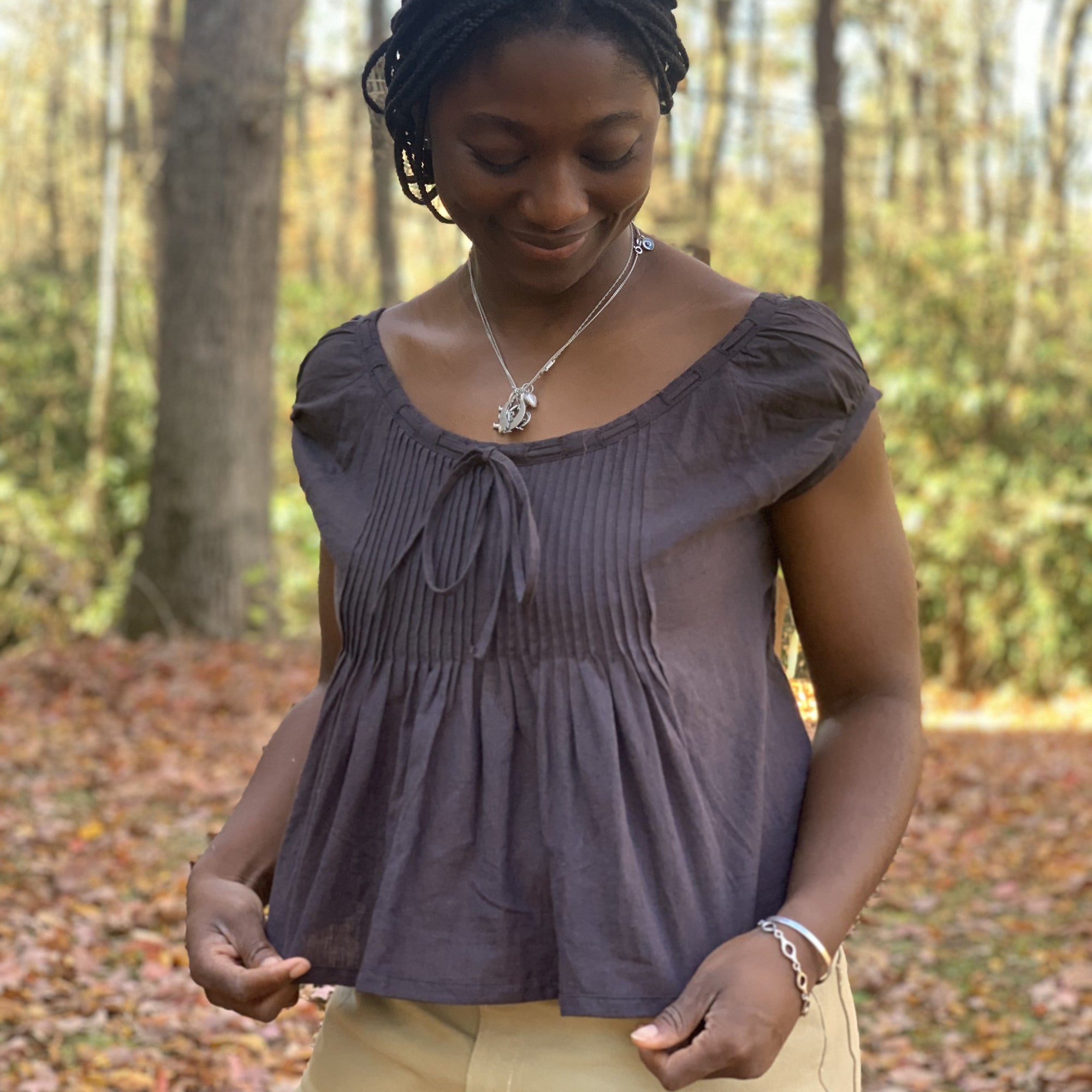 African American woman wearing 223 Chemise Blouse looking  down at the garment and head turned to her right. The top is made out of a cotton linen chambray in plum. she is outside with the orange, yellow and red leaves of fall on the ground.