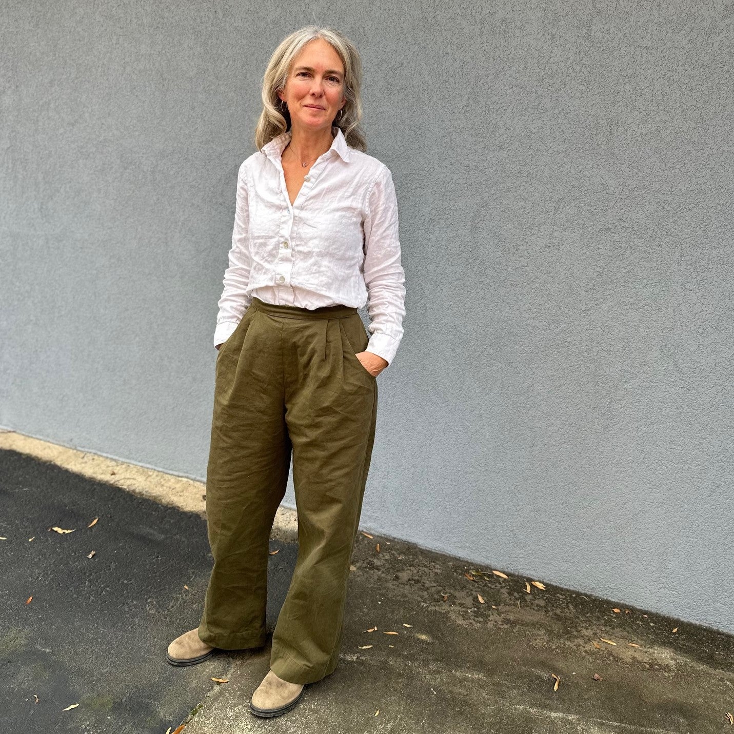 Woman standing in front of a grey wall wearing a white linen shirt and olive wide-legged trousers.