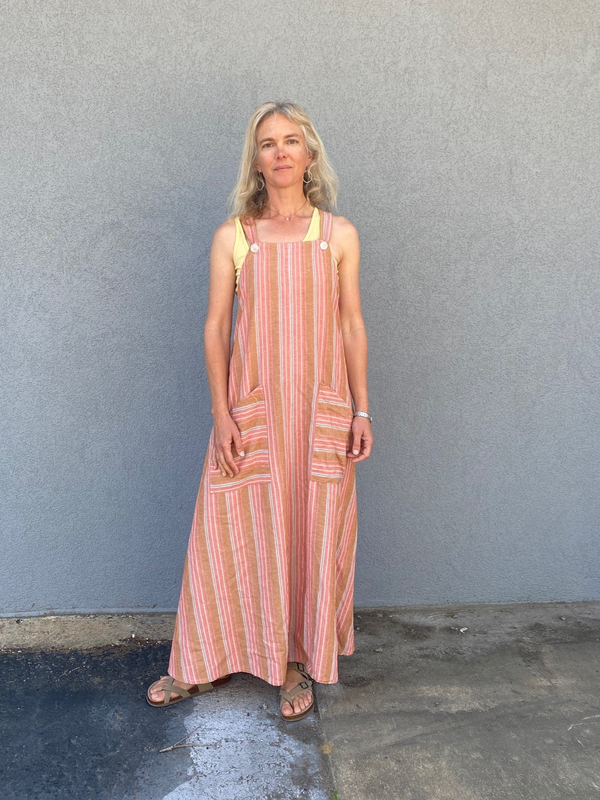 woman standing in front of a grey wall wearing a peach colored striped long pinafore dress