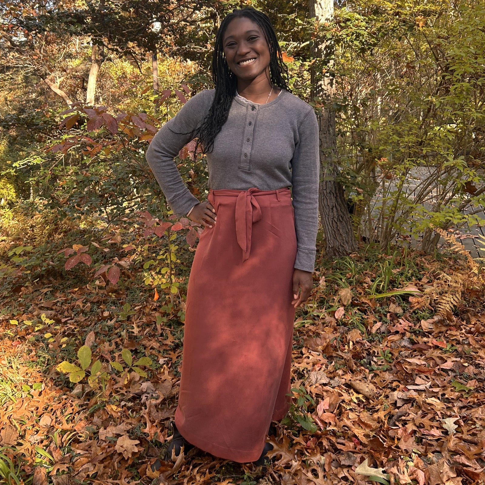 Woman standing and smiling with a hand on her hipoutside during autumn, wearing a grey long sleeved and the 131 Tibetan Chupa Wrap Skirt in the color auburn.