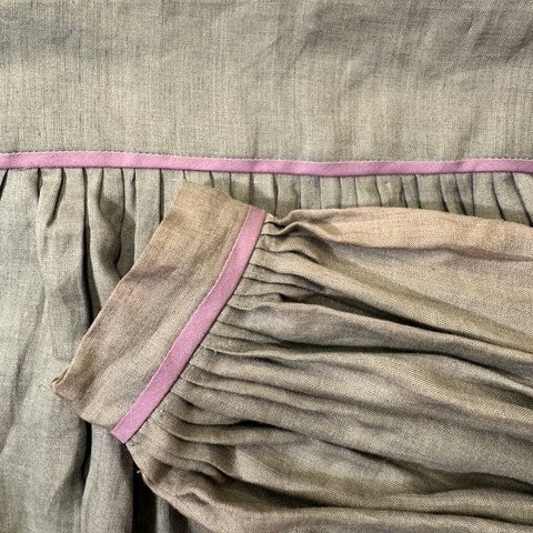 close up of a olive green blouse with many small pleats at the bodice and cuff, and with pink trim on the edges.