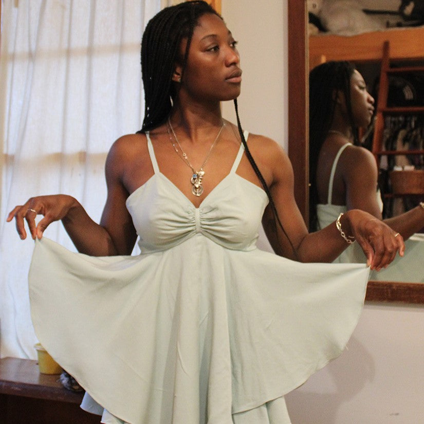  African American woman looking to her left, wearing a aqua light blue Juliette's Dream garment, in a room with her reflection in a mirror on a wall on the right, holding layered skirt out with both hands.