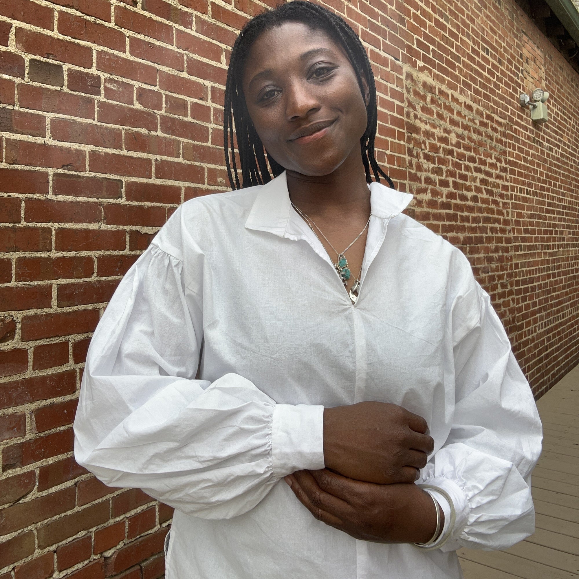 African woman wearing a white 147 Bunad blouse standing and smiling in front of a brick wall.
