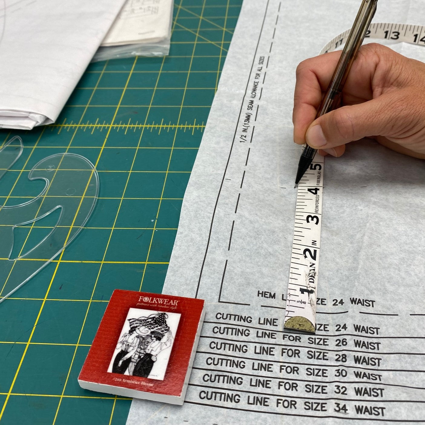 White paper pattern with a hand drawing a line near a white ruler.  Red paper weight on paper on a green mat.