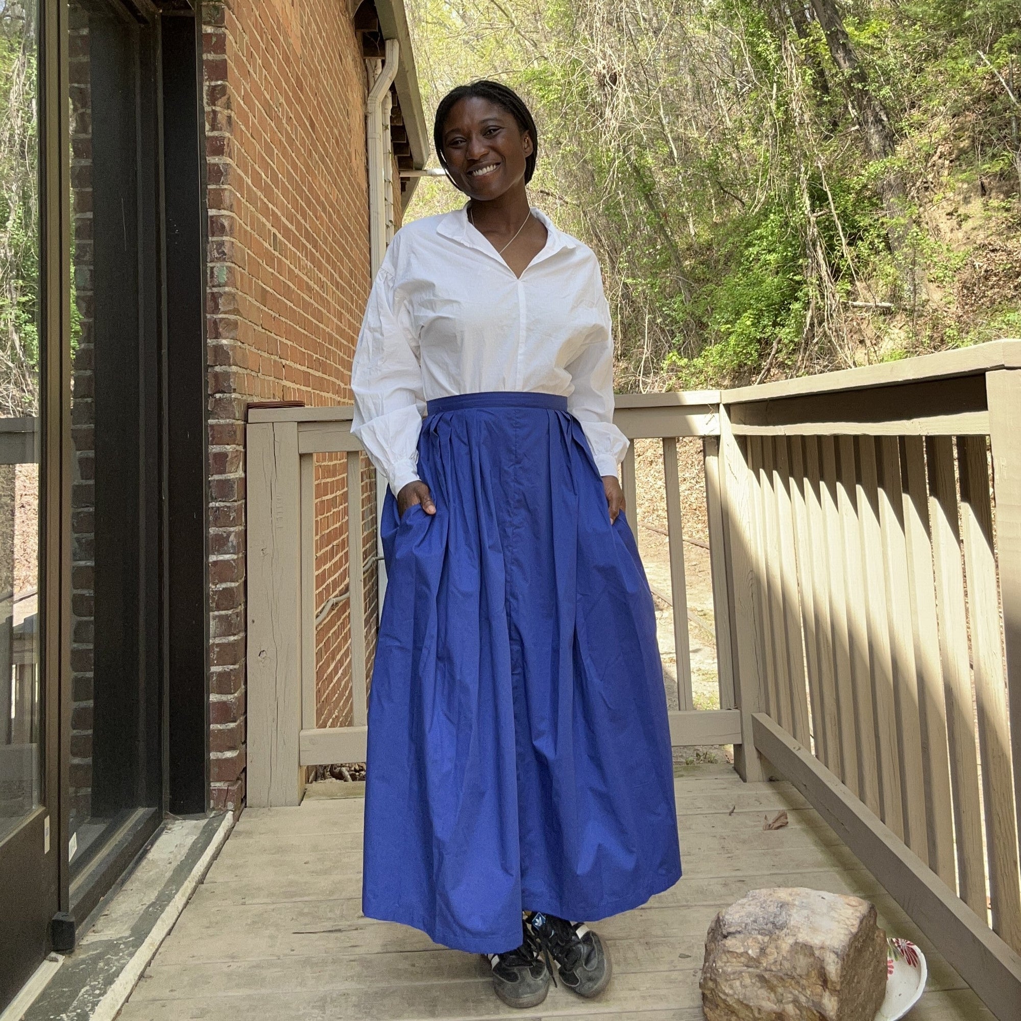 African woman wearing a white bunad blouse and royal blue pleated bunad skirt standing outside in front of a brick building and smiling with hands in her pockets.