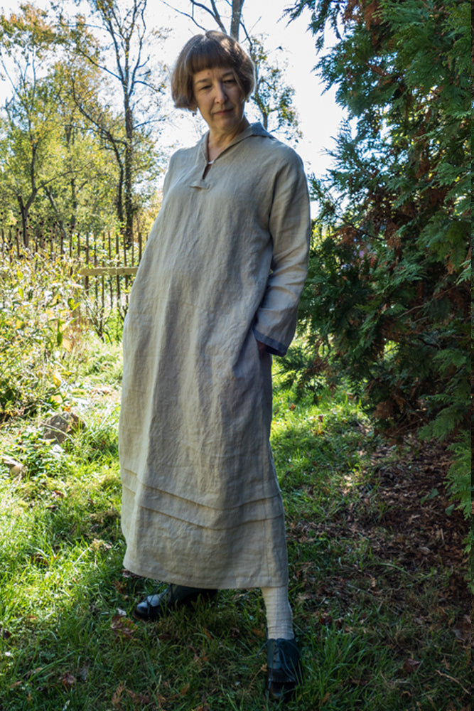 Woman standing in a garden wearing a long sleeve long sailor dress in grey linen.  Her hands are in her pockets.