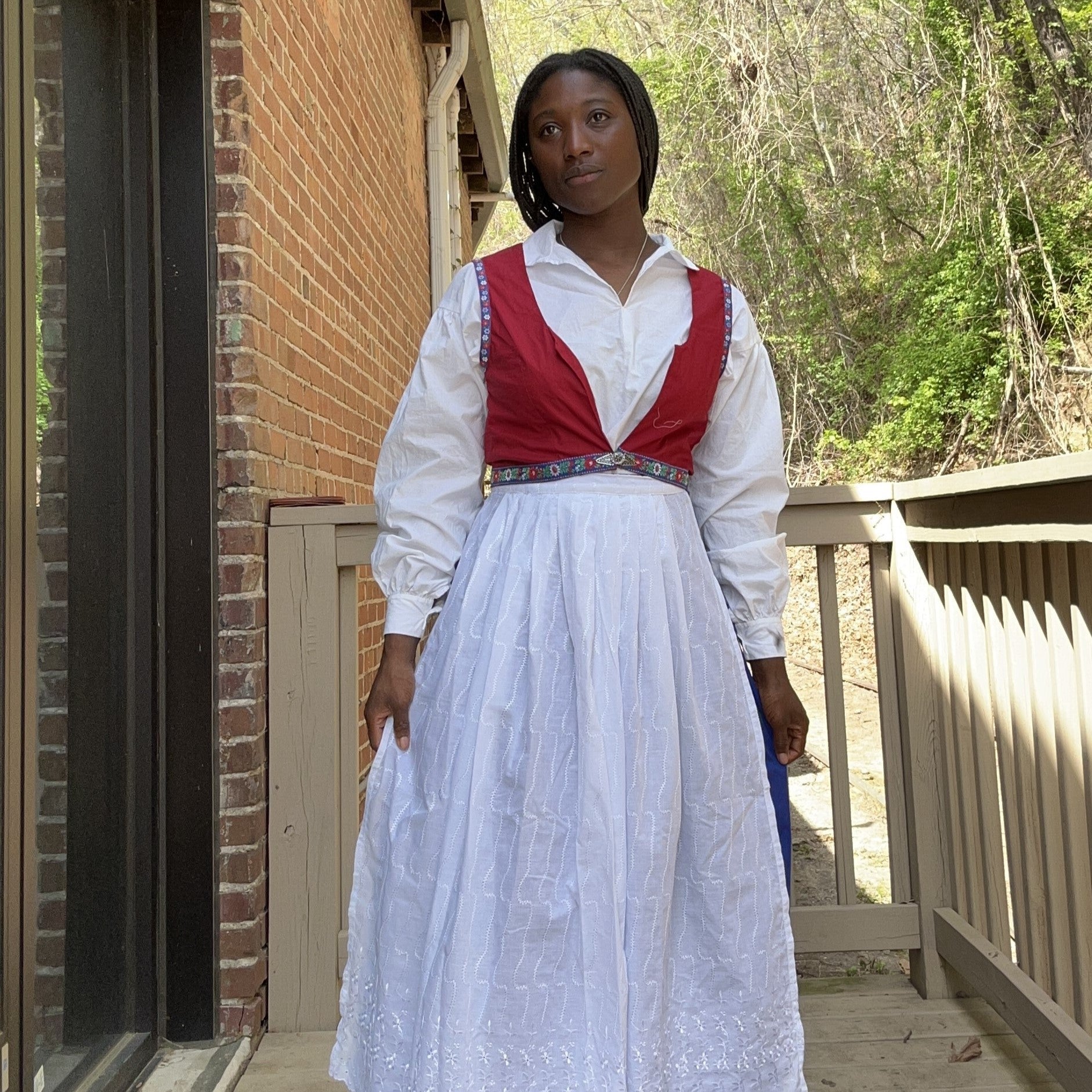 African woman wearing the 147 Bunad, White blouse, red vest with embroidery, royal blue skirt and white embroidered with silver thread apron. She is standing outside in front of a brick building with greenery behind her.