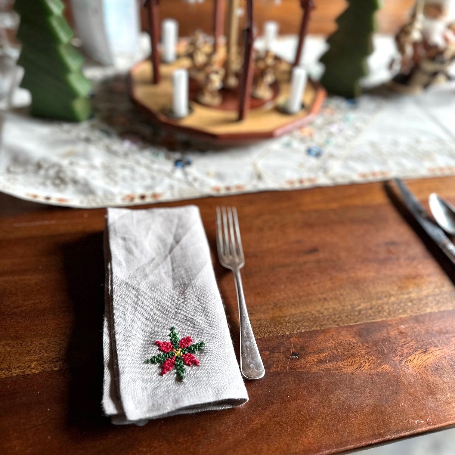 Napkin with a cross stitched poinsetta on a wooden table set with silverware and a table runner.