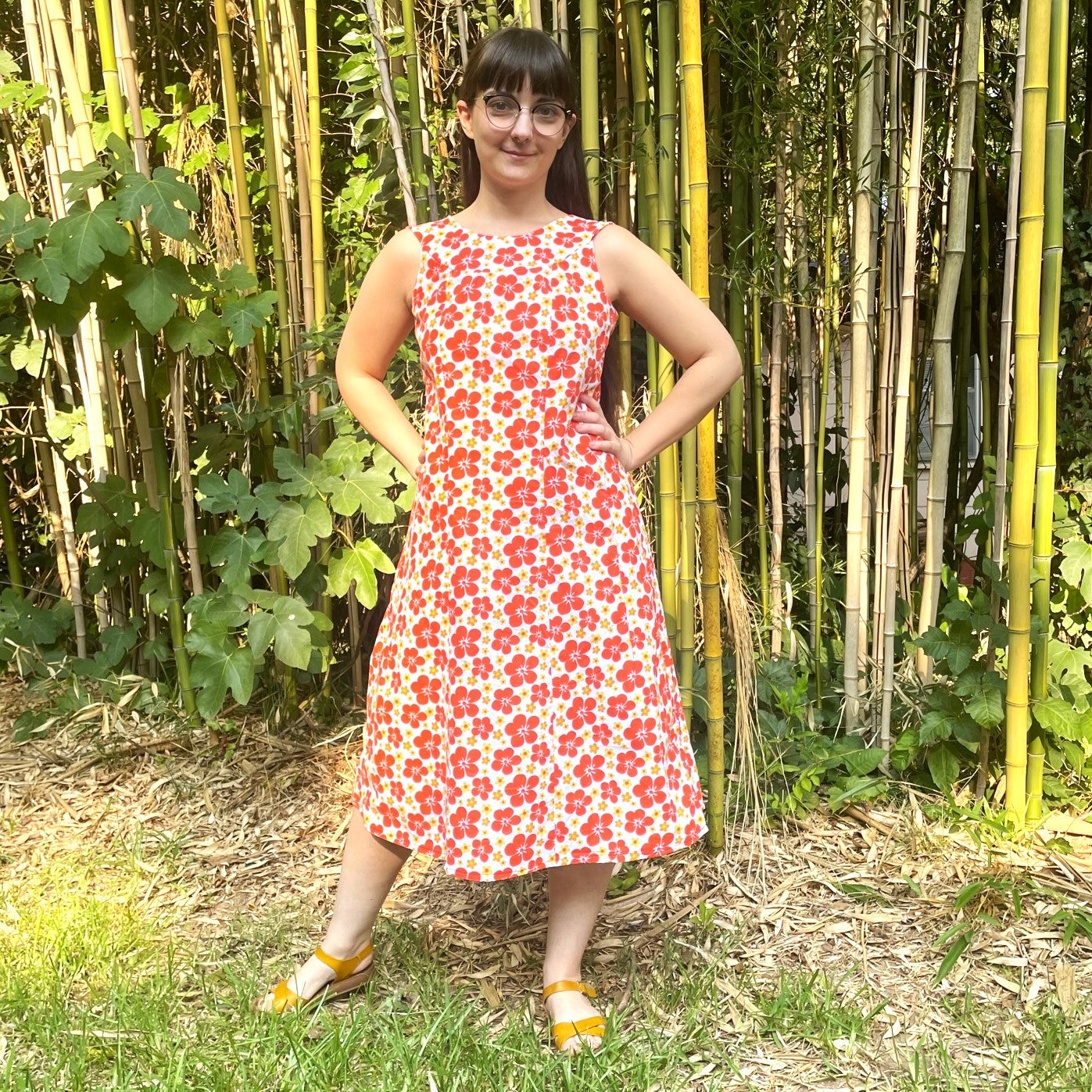 Woman standing with hands on hips in front of a bamboo forest, wearing an orange floral muumuu and yellow sandals.