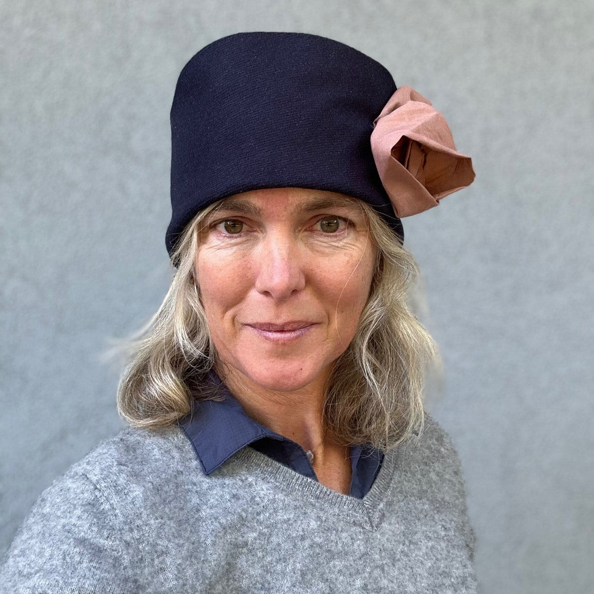 Photo of woman's from shoulders up wearing a grey sweater and navy cloche hat with a  pink flower