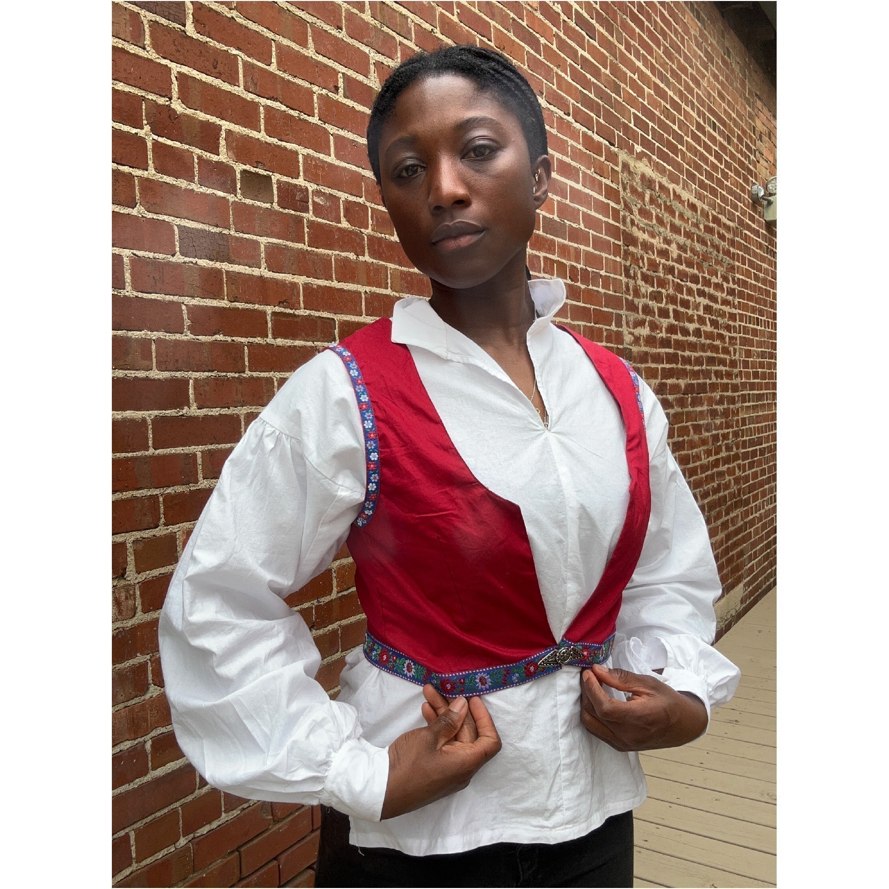 African woman wearing 147 white bunad blouse and red vest standing in front of a bring wall. With hands raised to the hem of the vest.