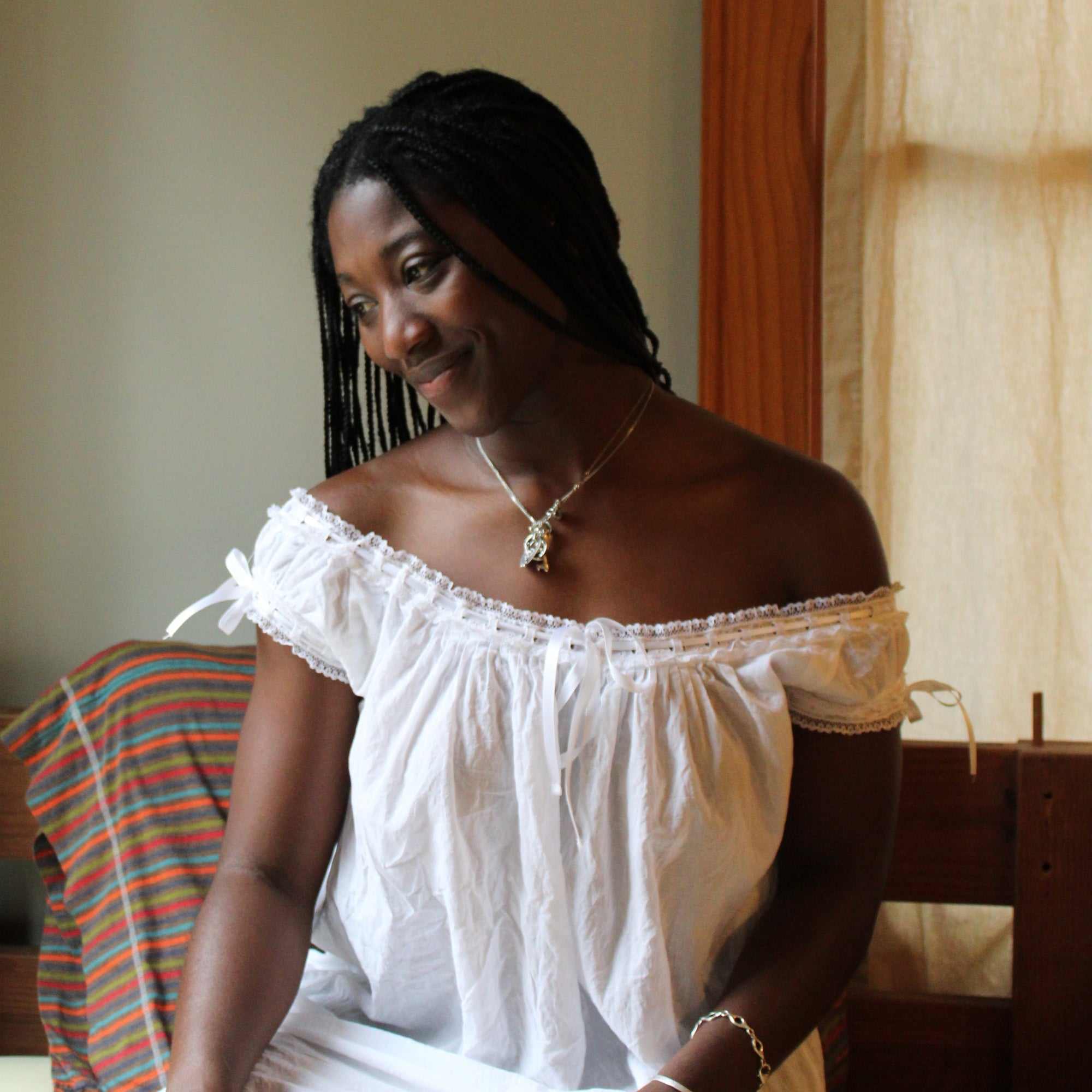 African american woman sitting on a bed with yellow sheets, wearing 223 A Lady's Chemise in white. Smiling down to her right.