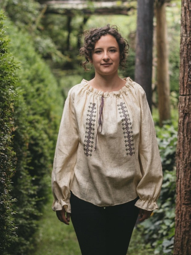Woman walking toward camera wearing a cream colored Romanian Blouse with ruffles at the cuffs and a gathered neckline.