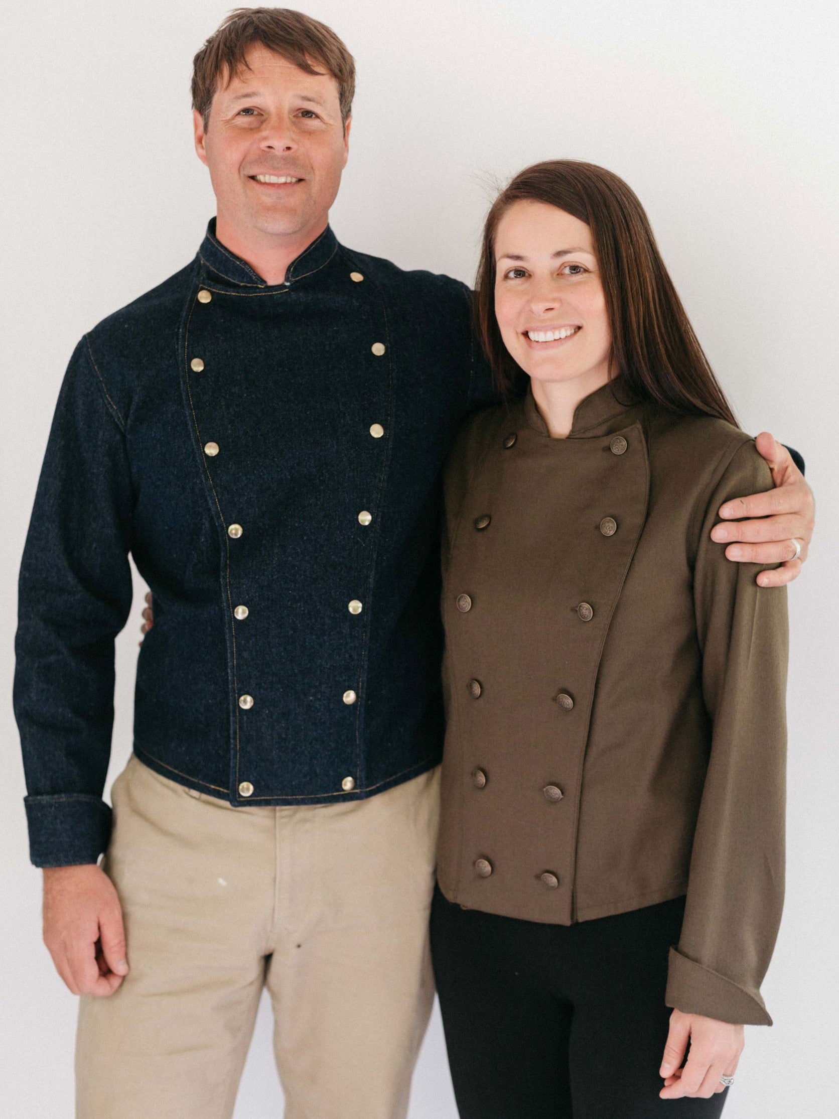 Man and woman wearing Belgian Military Chef's Jackets.  Woman on the right has on an olive coat.  Man on left has a dark blue denim coat.  Coats are double breasted with two rows of buttons.  Man's arm is around the woman's shoulders.