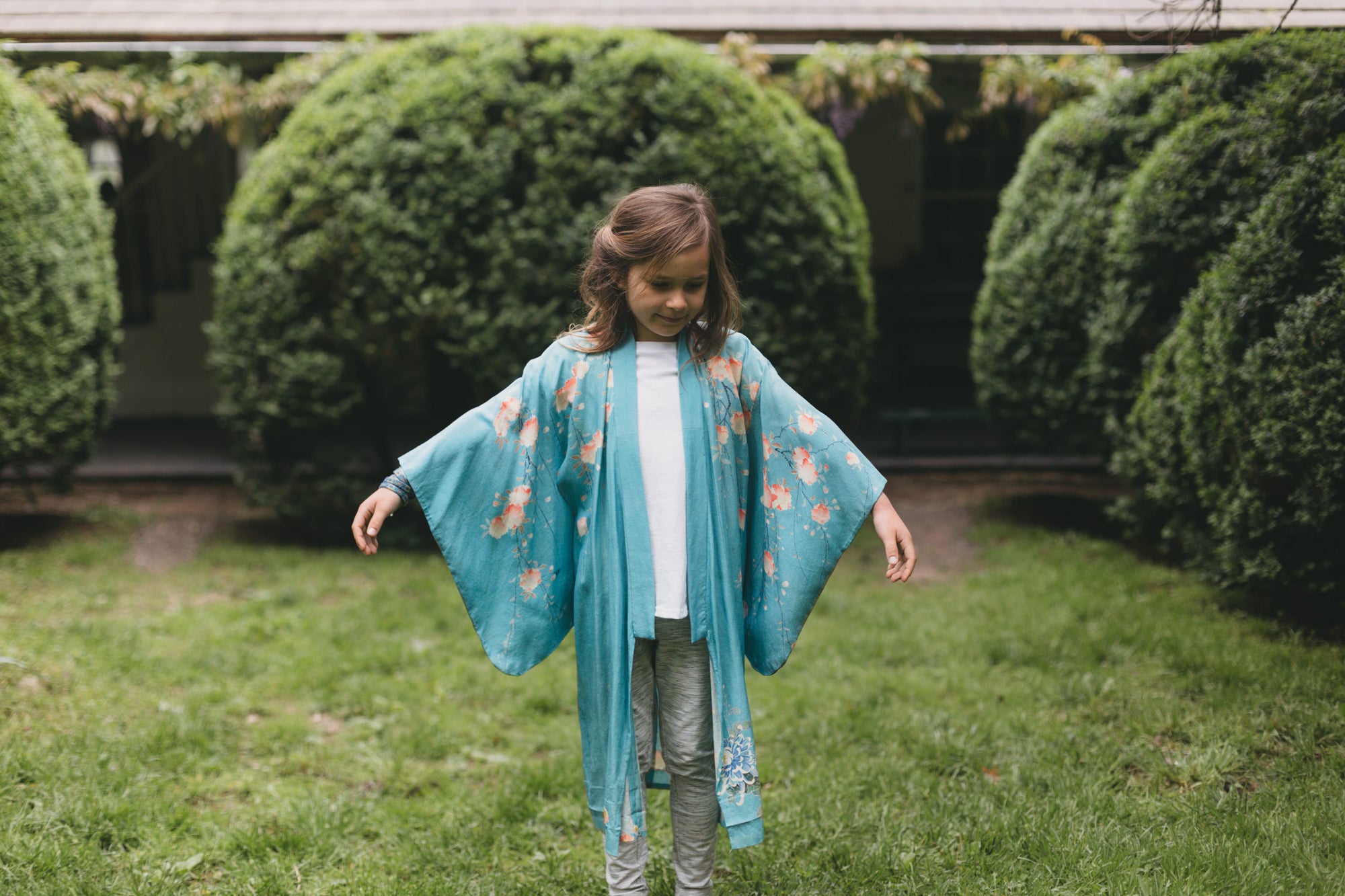 Young girl wearing a blue floral kimono standing in a garden with manicured hedges.