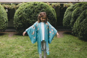 Young girl wearing a blue floral kimono standing in a garden with manicured hedges.