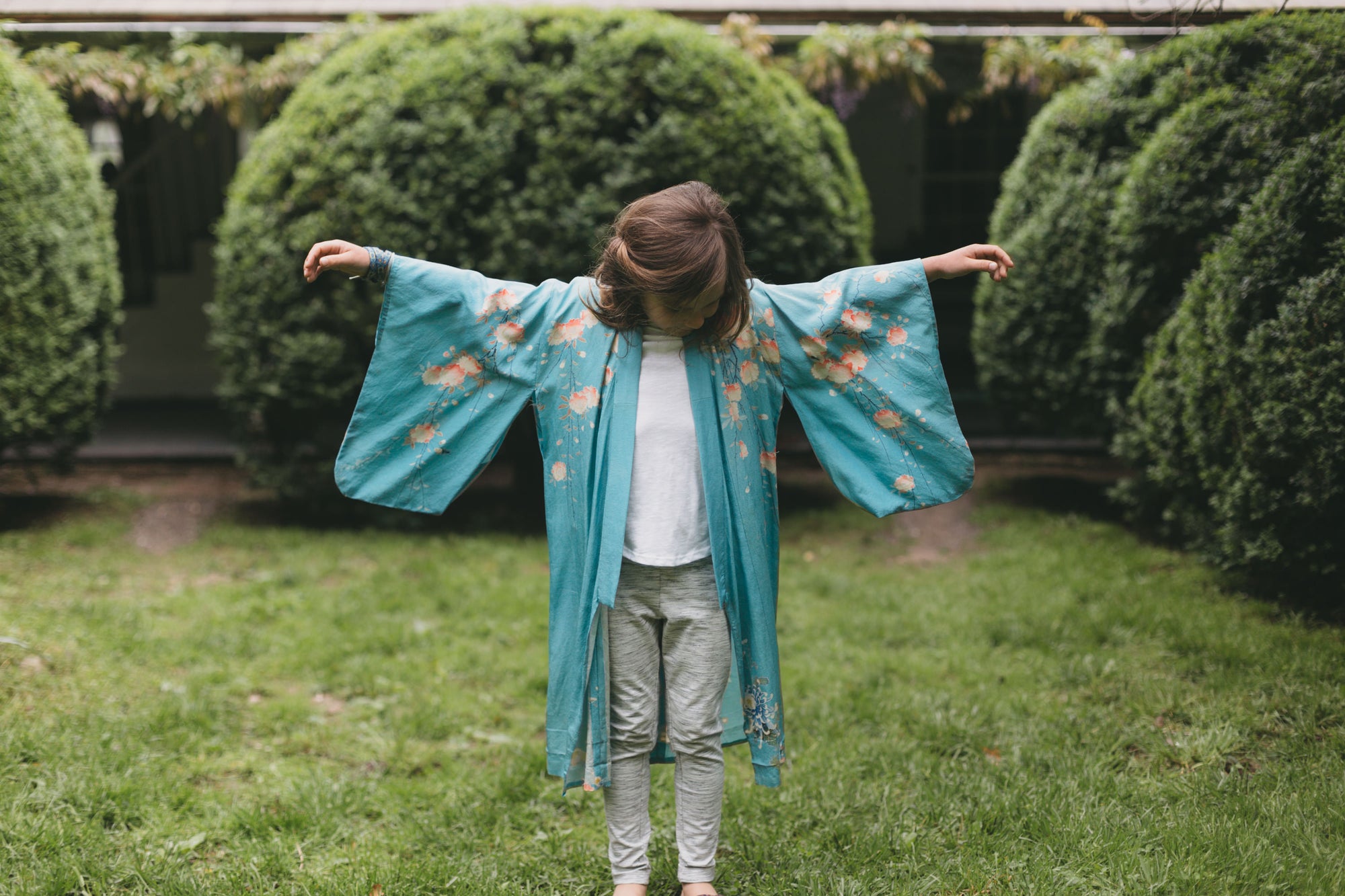 Child wearing a blue floral kimono standing in a garden with green bushes.