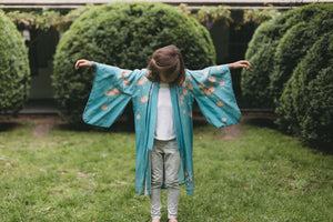Child wearing a blue floral kimono standing in a garden with green bushes.